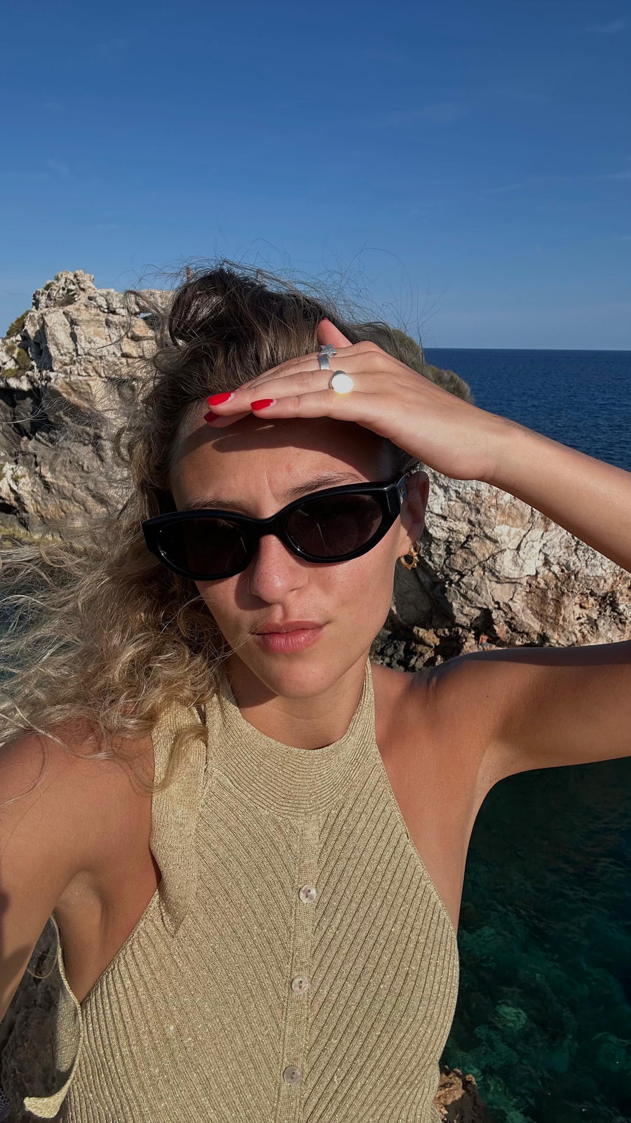 Woman with sunglasses and a ring, standing by a rocky coastline with blue sky and ocean.