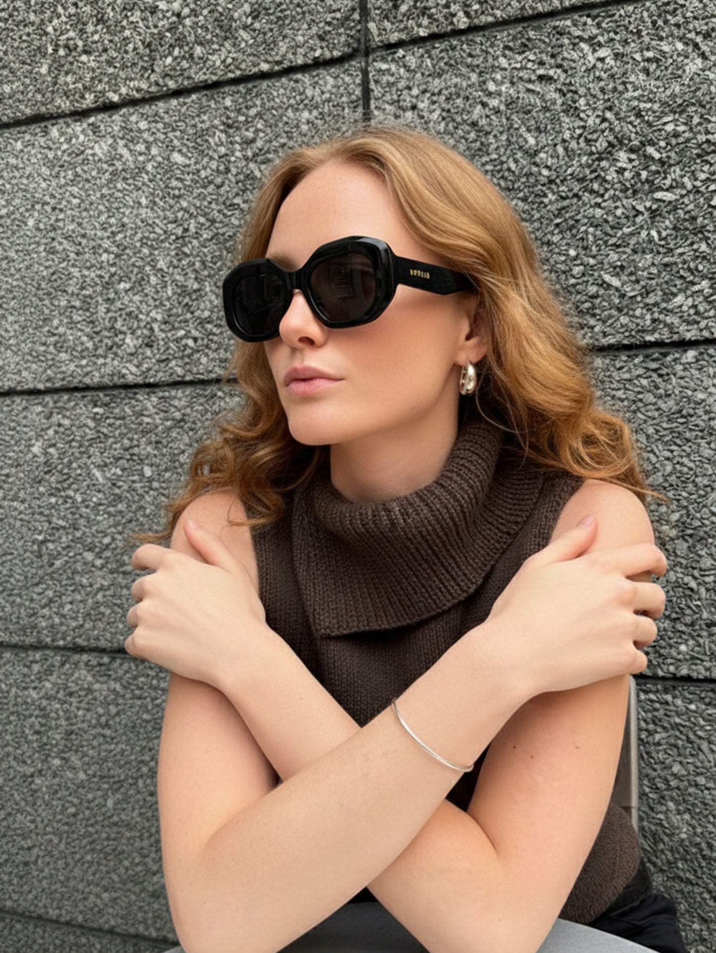 Woman wearing sunglasses and a brown sleeveless top against a gray brick wall