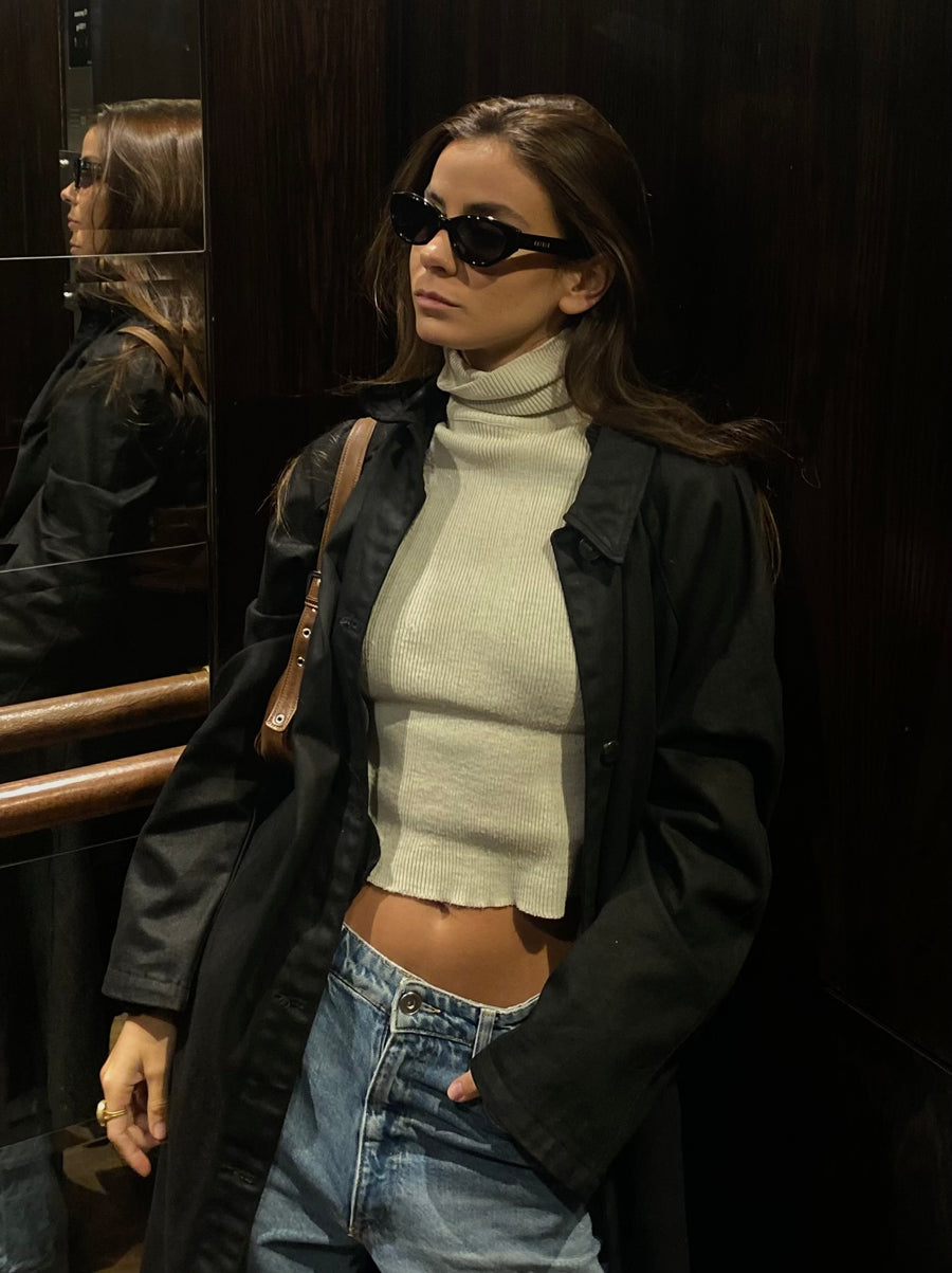 Woman wearing sunglasses, a black jacket, white turtleneck, and blue jeans in an elevator.