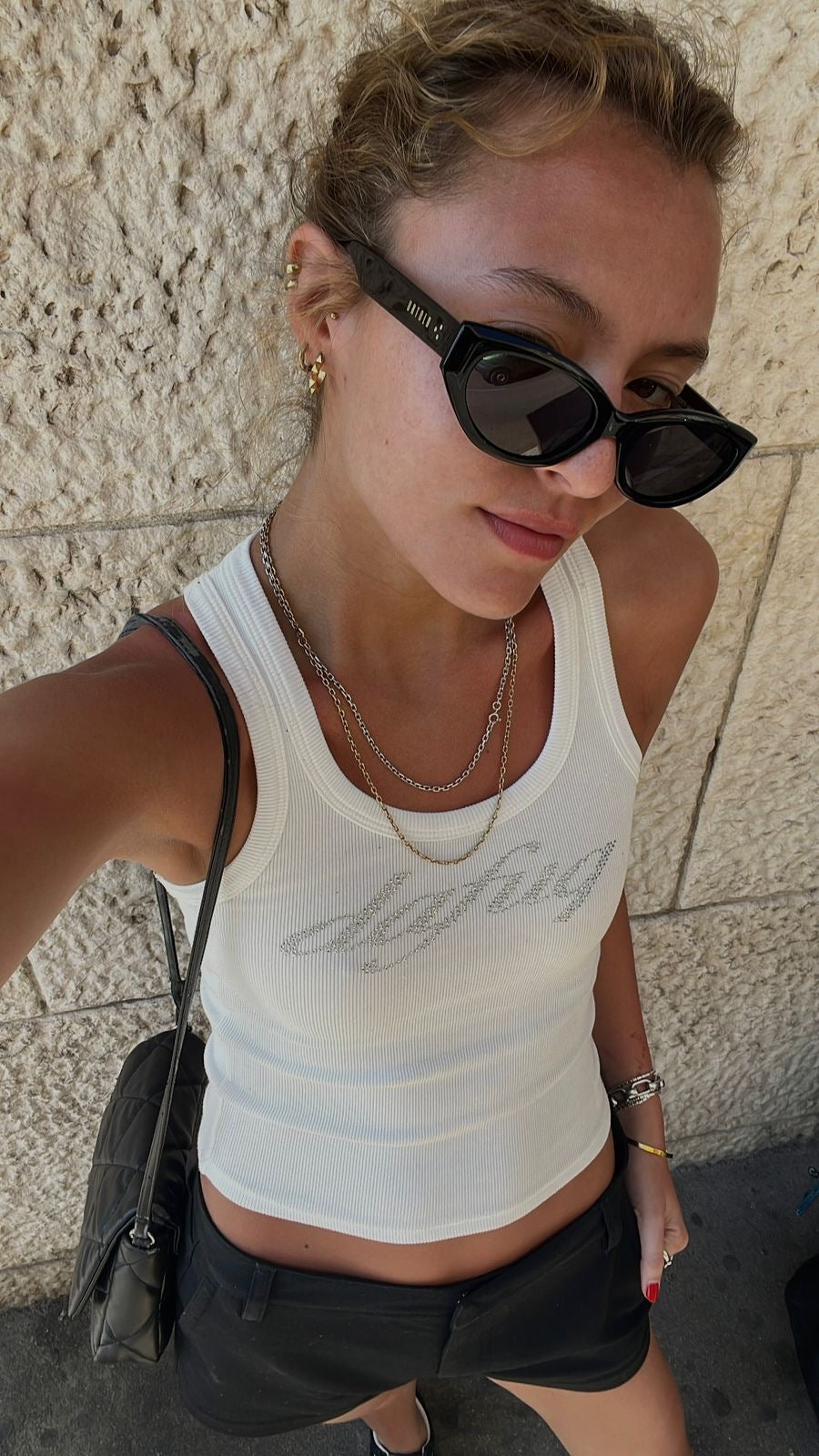 Woman wearing a white tank top and black sunglasses against a stone wall.