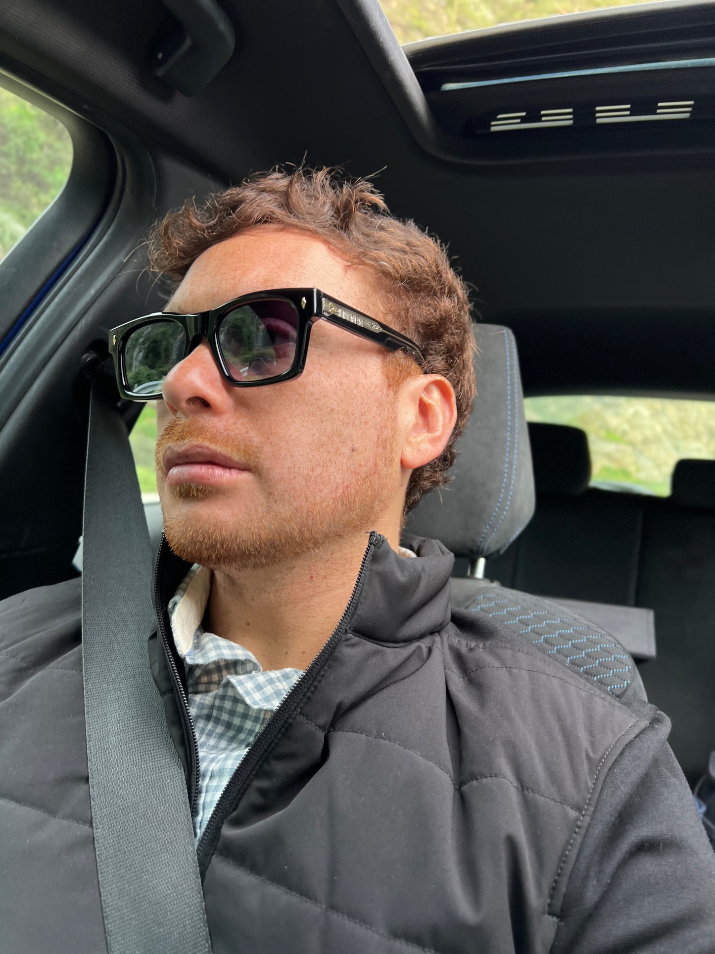 Man wearing sunglasses and a gray jacket sitting in a car.
